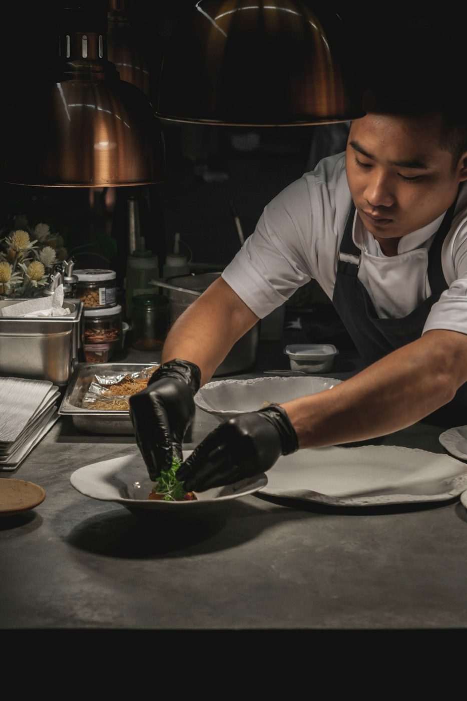 A chef in a white coat and black gloves carefully plating a garnished dish under kitchen heat lamps