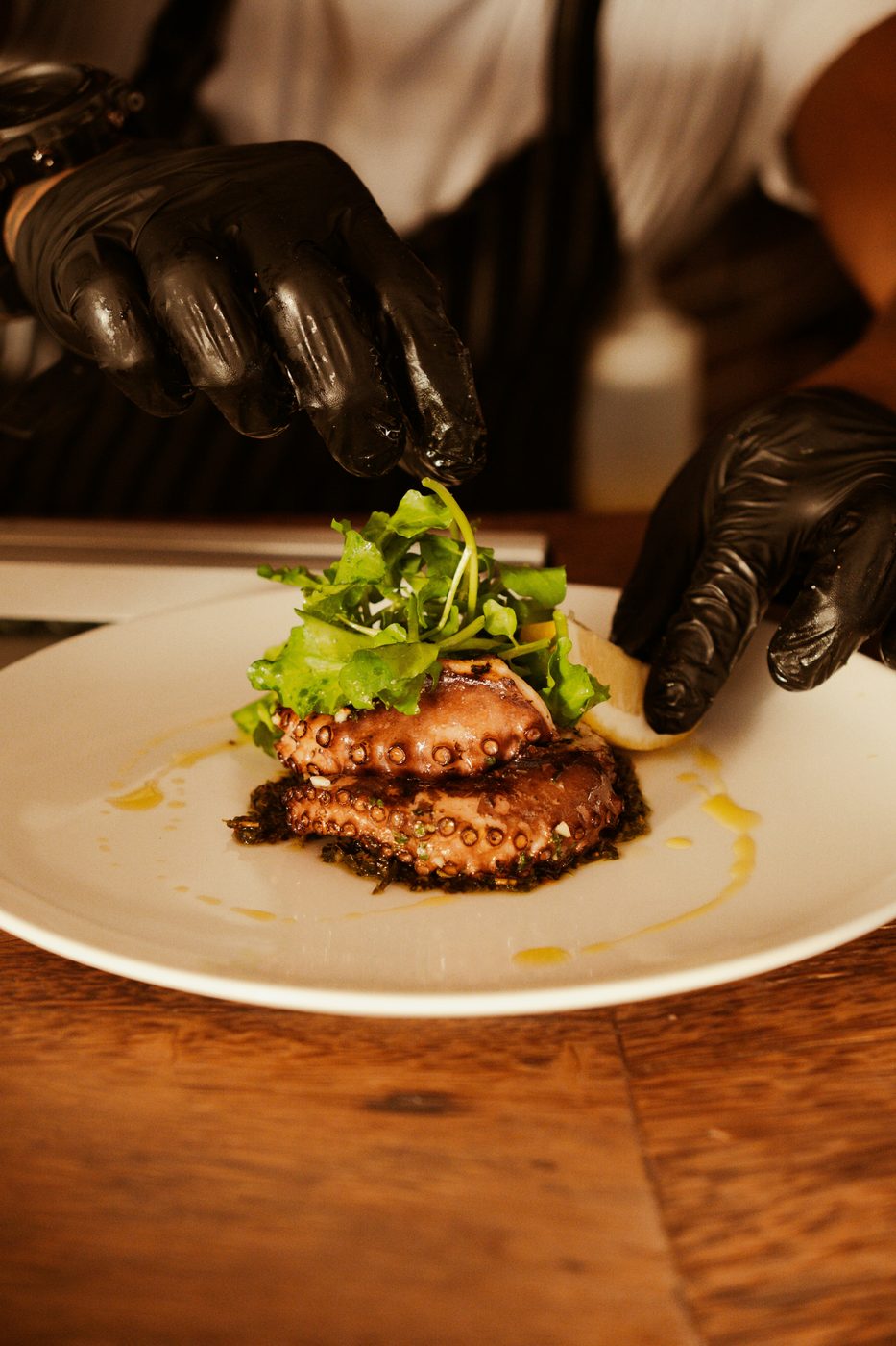 A chef in black gloves plating grilled octopus with greens on a white plate