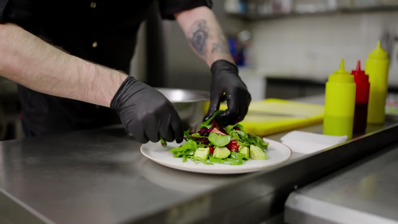 Gloved hands plating a salad at a stainless-steel pass in a commercial kitchen