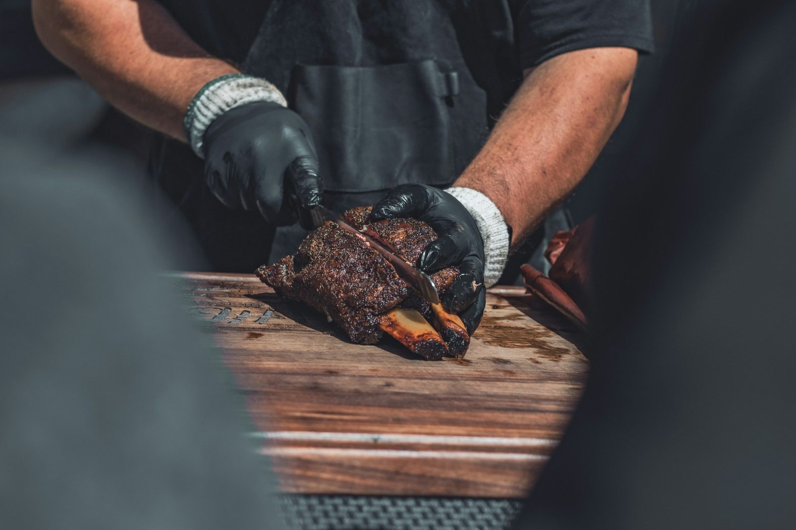 A cook wearing black nitrile gloves slicing smoked beef ribs on a wooden cutting board